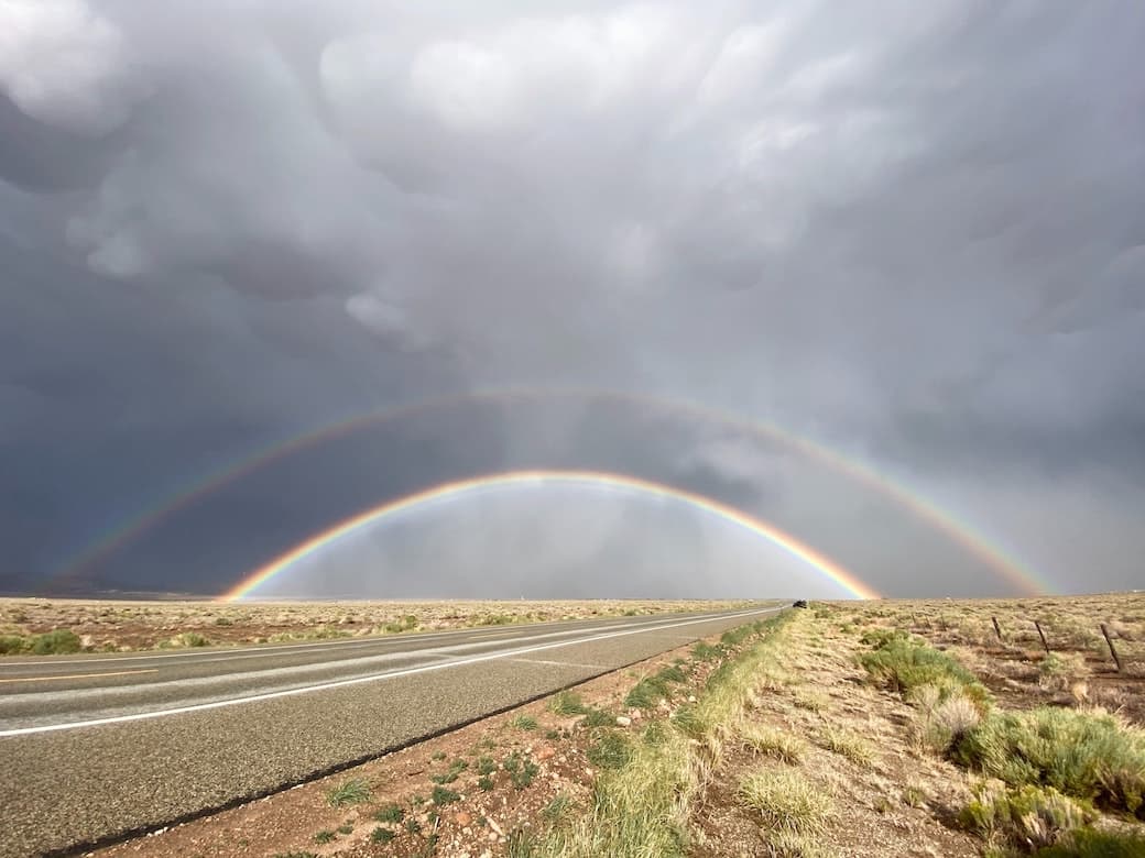 Photo of a flat landscape with a road in the foreground in cloudy weather with a double rainbow visible.