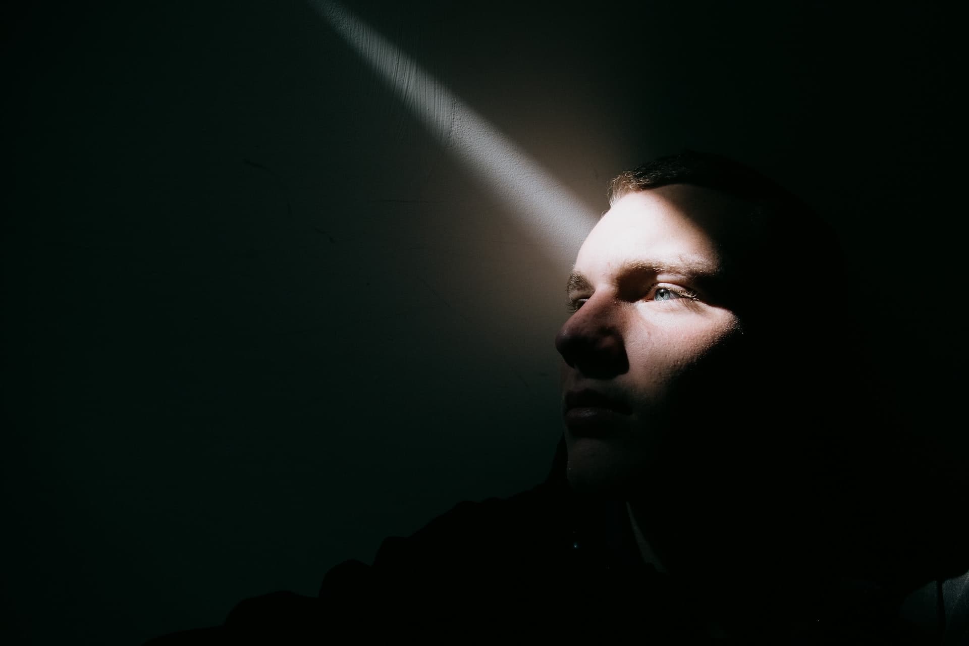 A man in a dark setting with a single spotlight shining into his left eye illuminating part of his face.