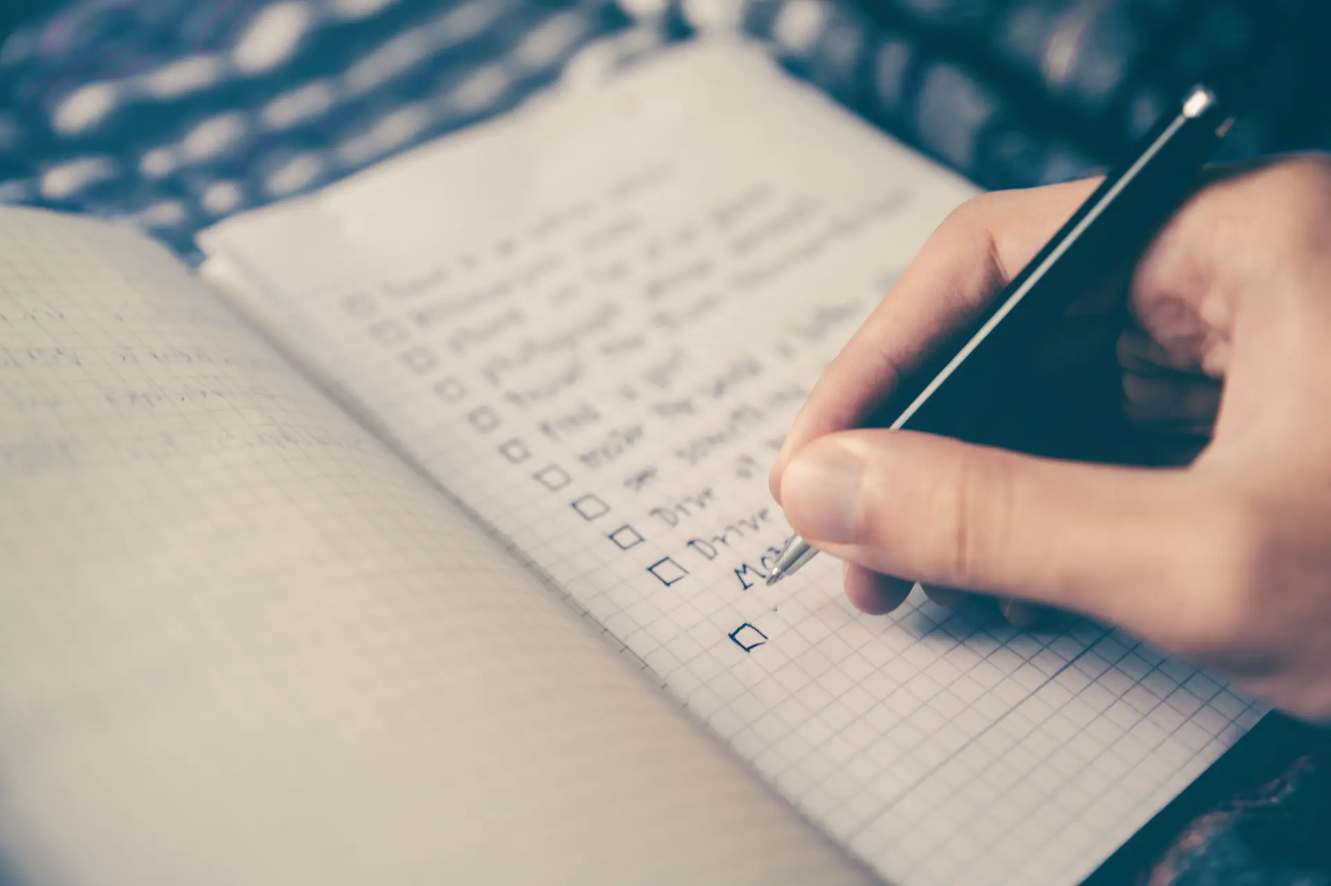 A notebook with grid pattern with hand written checklist items being written on it.