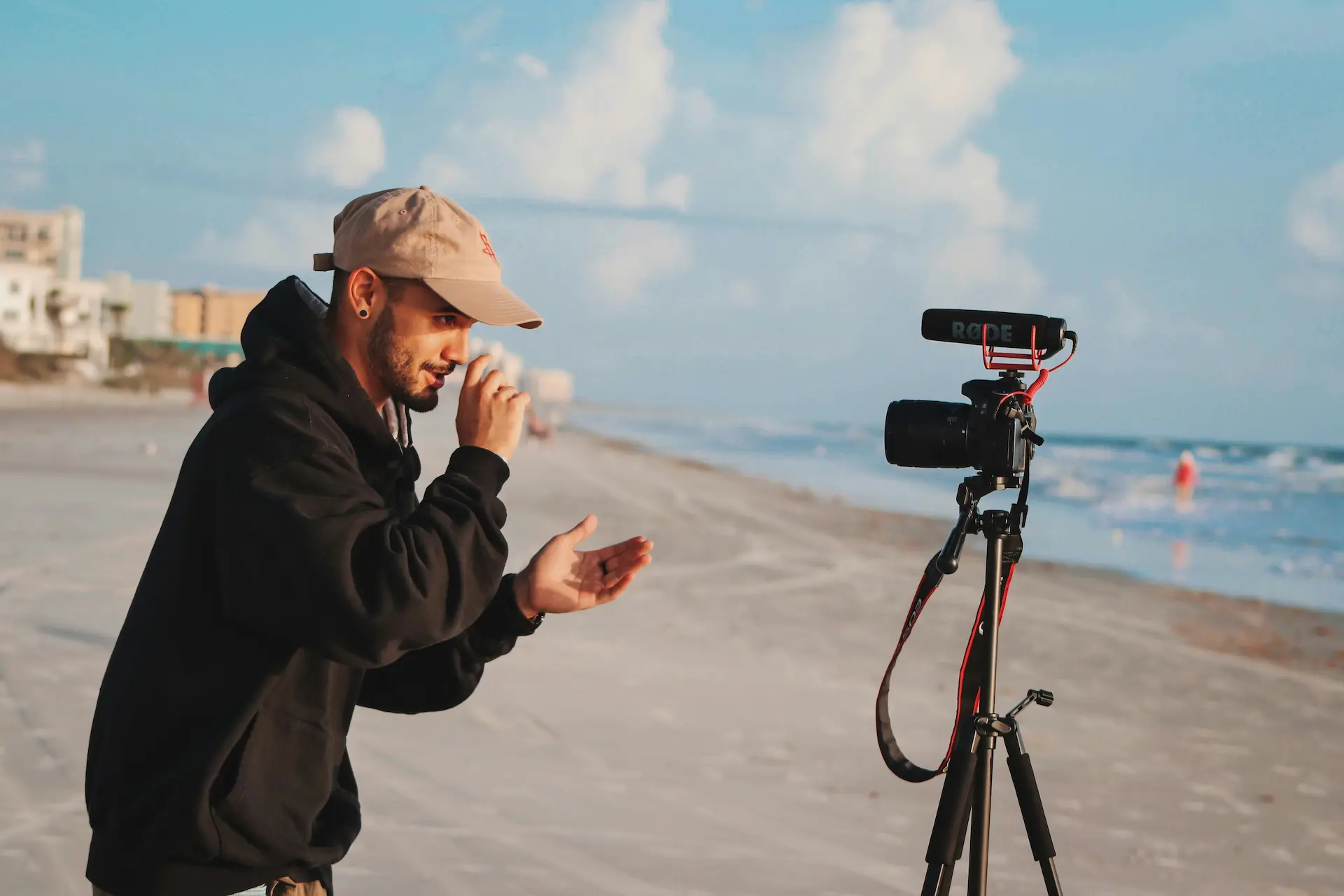 White guy on beach looking into a camera with a microphone on a tripod explaining something for a recording.