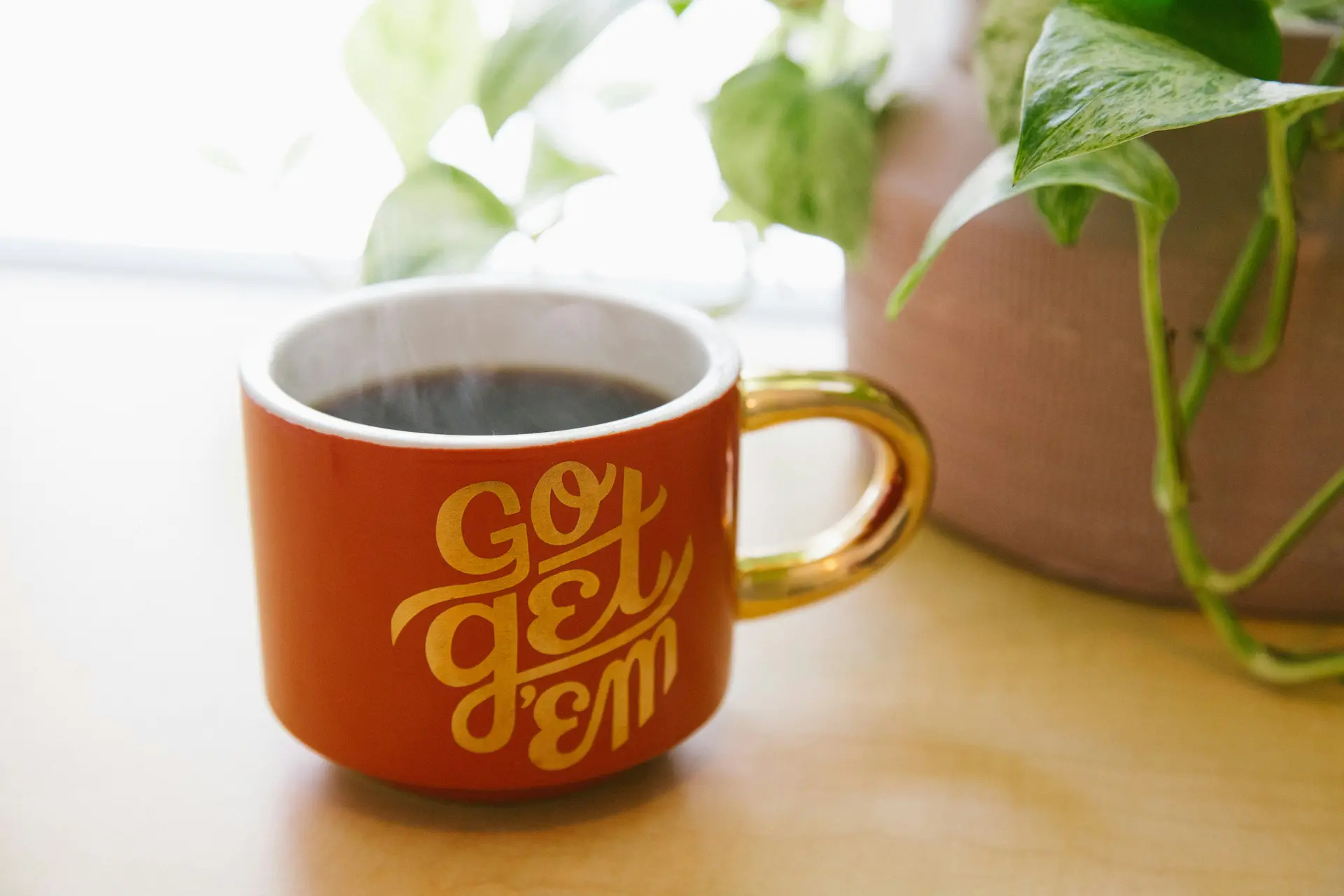 Red mug with yellow writing on it that says go get'em! It has steaming coffee in it. Pot of plant next to it.