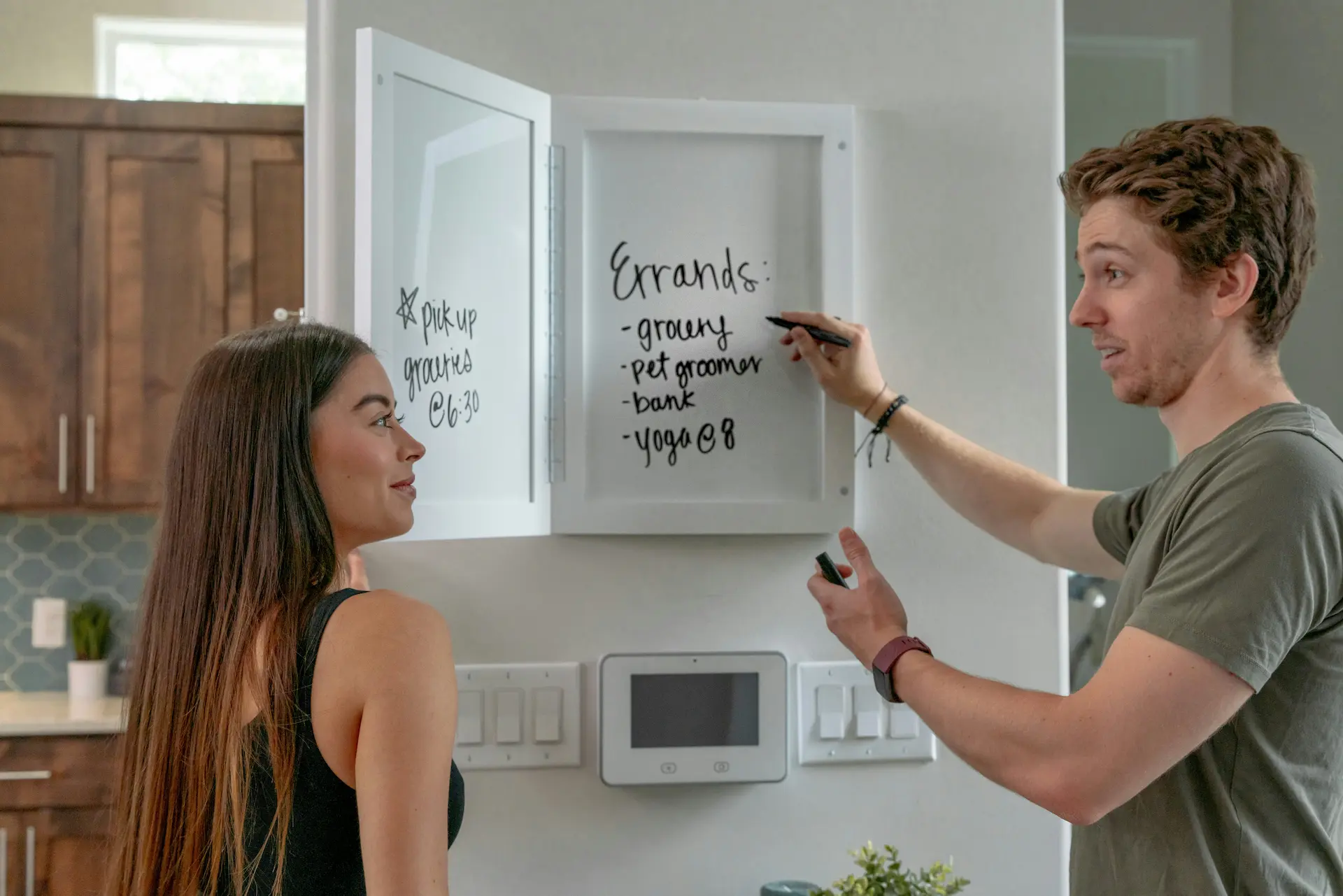 Couple standing in their kitchen looking at a wall mounted whiteboard they use as a todo list.