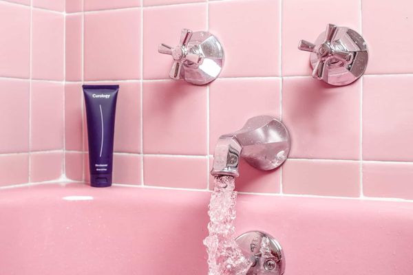 A close up photo of a tap and two knobs of a very pink bathtub in a very pink tiled bathroom. Water flows out of the tap.