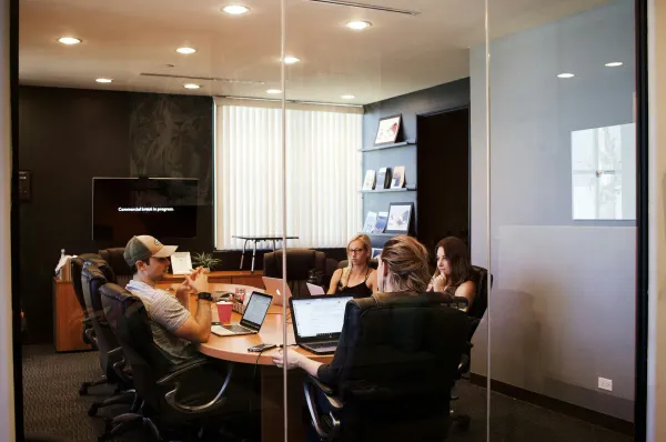 Photo of a meeting room in a corporate office behind glass walls. Four people are sitting at a table with laptops.