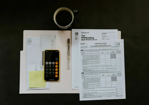 Folio of paper tax forms, cup of coffee, a pen, and the calculator app on an iPhone.