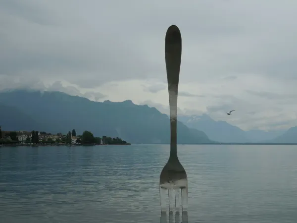 A huge fork statue that's stabbing a lake with mountains in the background.