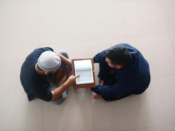 A teacher explaining something in the Quran to a student sitting opposite. Between them the open Quran on a tiny table. The photo was shot from above.